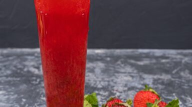 A refreshing strawberry cocktail garnished with a whole strawberry and mint leaves on a wooden coaster, surrounded by fresh strawberries, perfect for juicing with strawberries.