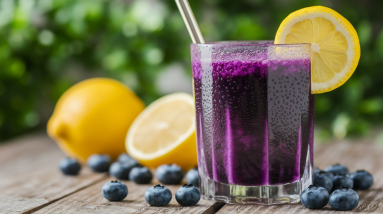 A glass of purple blueberry lemon juice with a lemon slice on the rim and a metal straw sits on a wooden surface, surrounded by fresh blueberries and whole lemons—a vibrant antioxidant juice for a natural health boost.