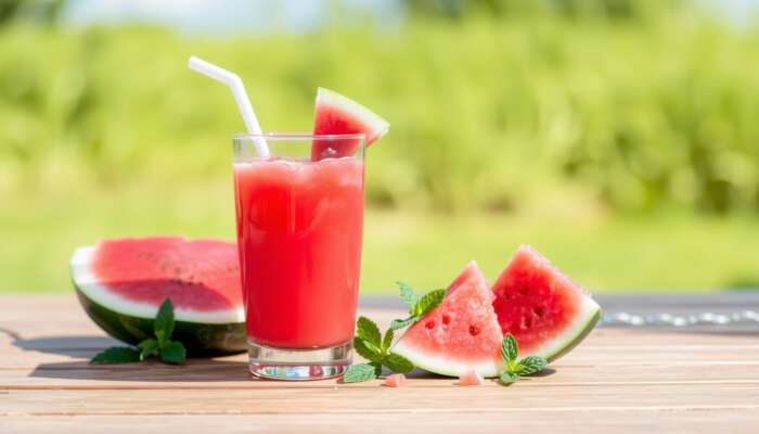 A glass of bright pink watermelon juice surrounded by fresh watermelon chunks and mint leaves on a sunny picnic table with a blurred green grass and blue sky background.